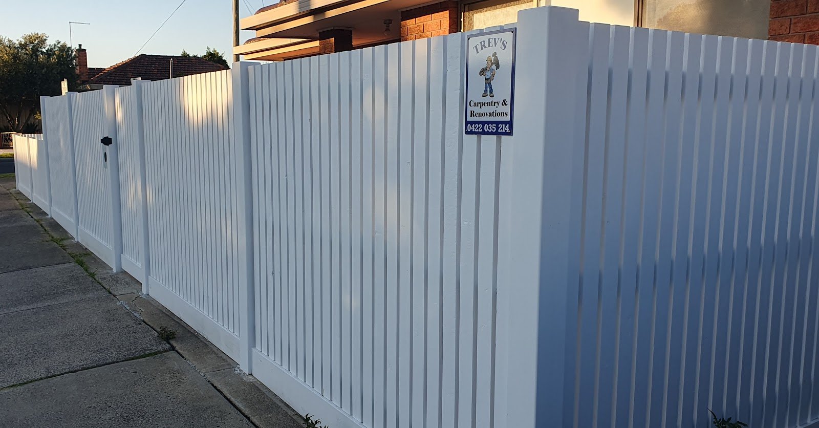 Corner of a white picket fence with Trev's Carpentry & Renovations sign attached